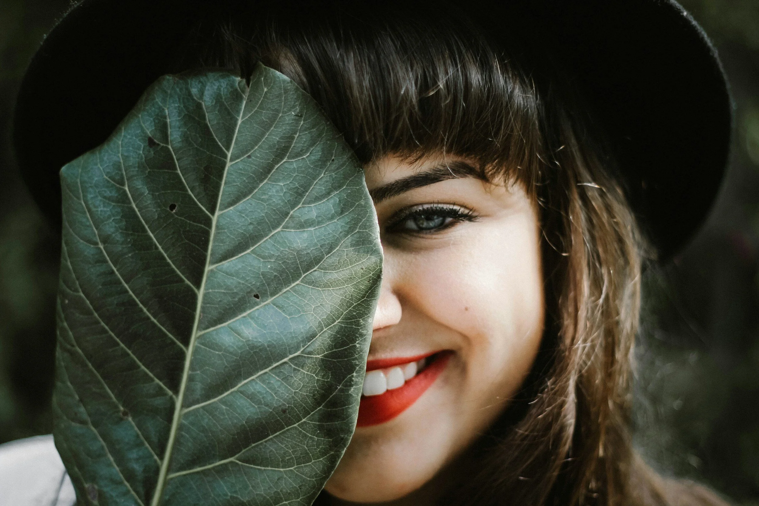 Photo of a pretty girl with dark hair and red lipstic, hiding half of her face with a pretty, thick leaf.