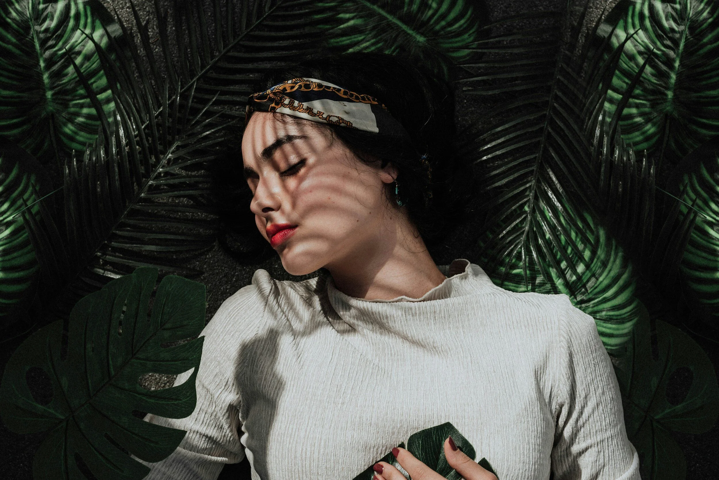 Photo of a pretty dark haired girl with white lipstic laying on top of dark green palm leaves.